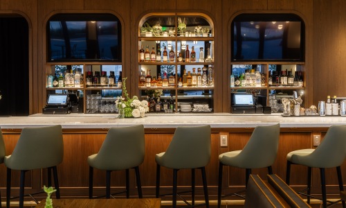 Inside Mila's restaurant at Gibson Point with modern seating and upscale finishes Elegant interior of Mila’s bar at Gibson Point, showcasing stylish green barstools, marble countertops, floral arrangements, and a curated selection of spirits in a warm wood-paneled setting.